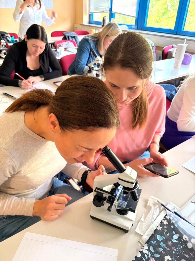 Zwei Frauen betrachten gemeinsam eine Probe durch ein Mikroskop in einem Klassenzimmer, weitere Personen arbeiten im Hintergrund.