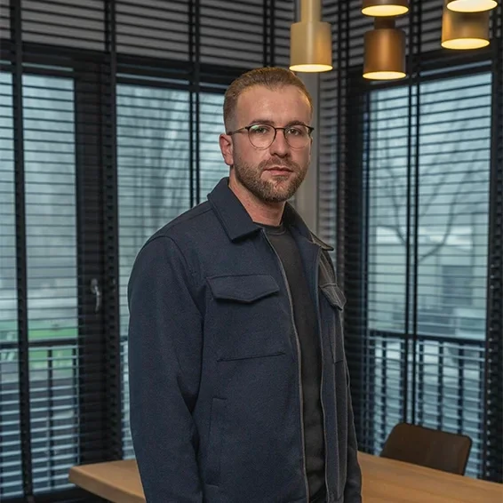 Astrit Rrahmani steht in einem modernen Büro mit großen Fenstern und blickt in die Kamera.
