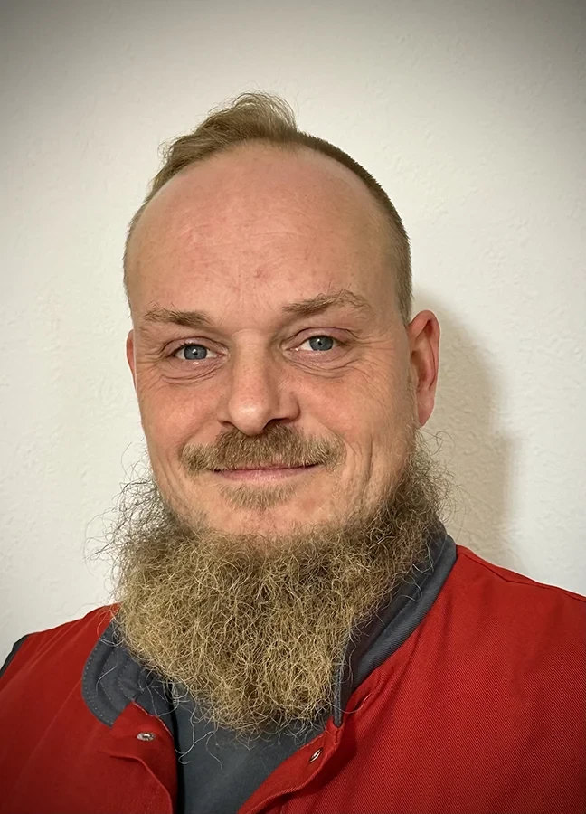 Aleksander Hein mit Bart und rotem Oberteil vor weißer Wand