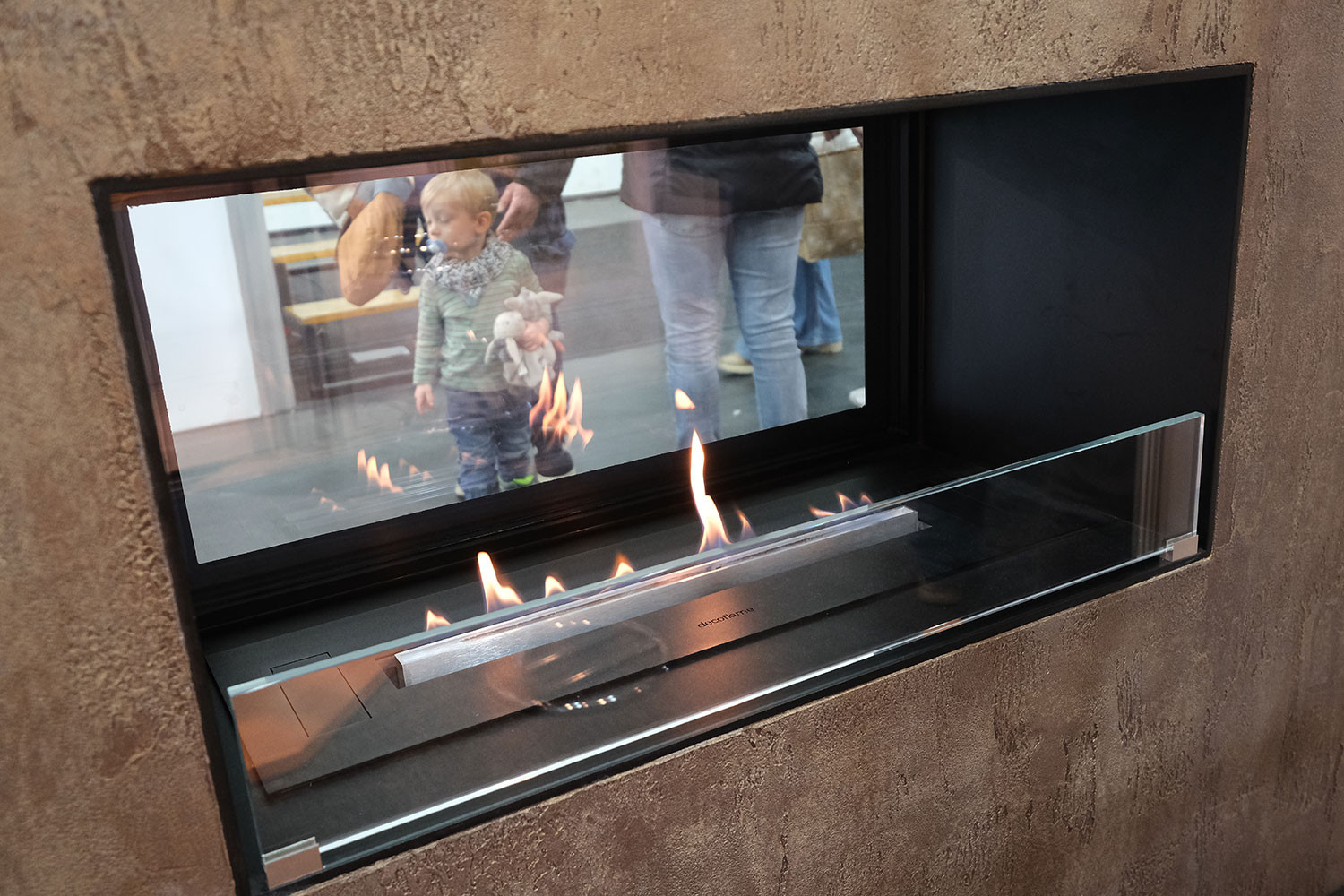 Kind mit Stofftier spiegelt sich in Glasscheibe eines modernen Kamins mit Flammen