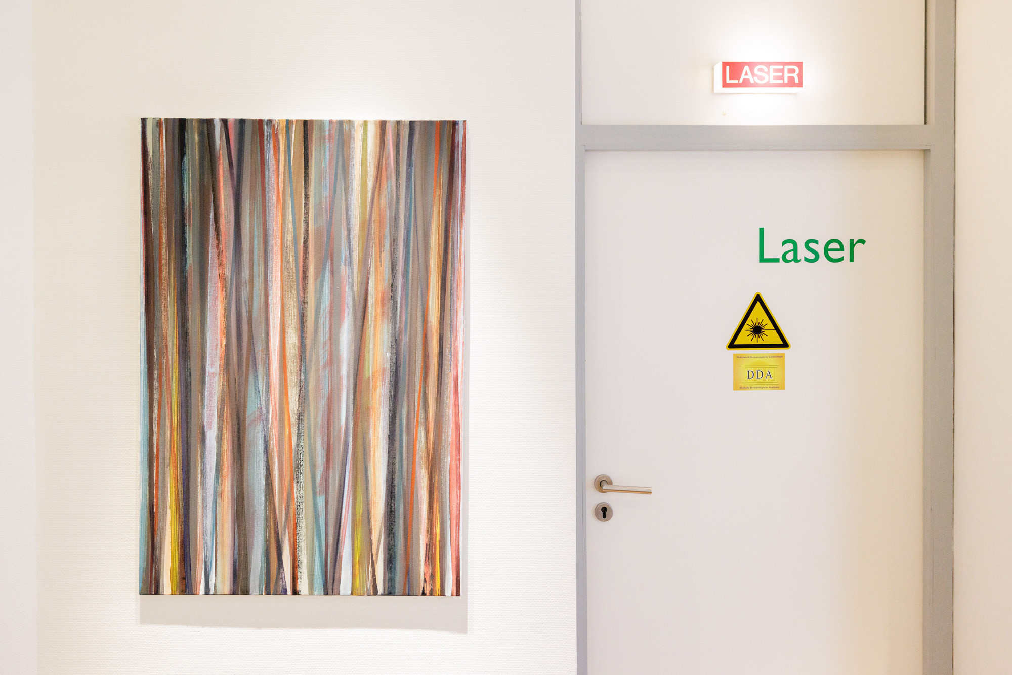 Praxisfoto 2025: Tür zum Laserraum mit Warnschild und moderner Kunst im Wartebereich