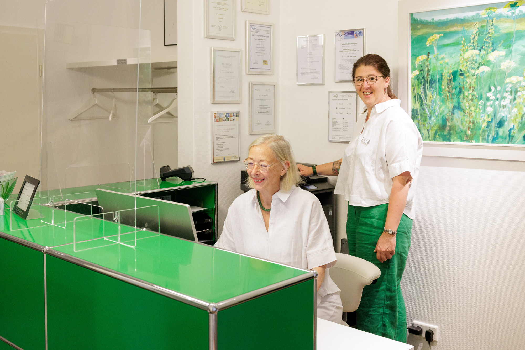 Empfangsbereich einer modernen Arztpraxis mit zwei Mitarbeiterinnen am Schreibtisch, grüne Theke und Zertifikaten an der Wand
