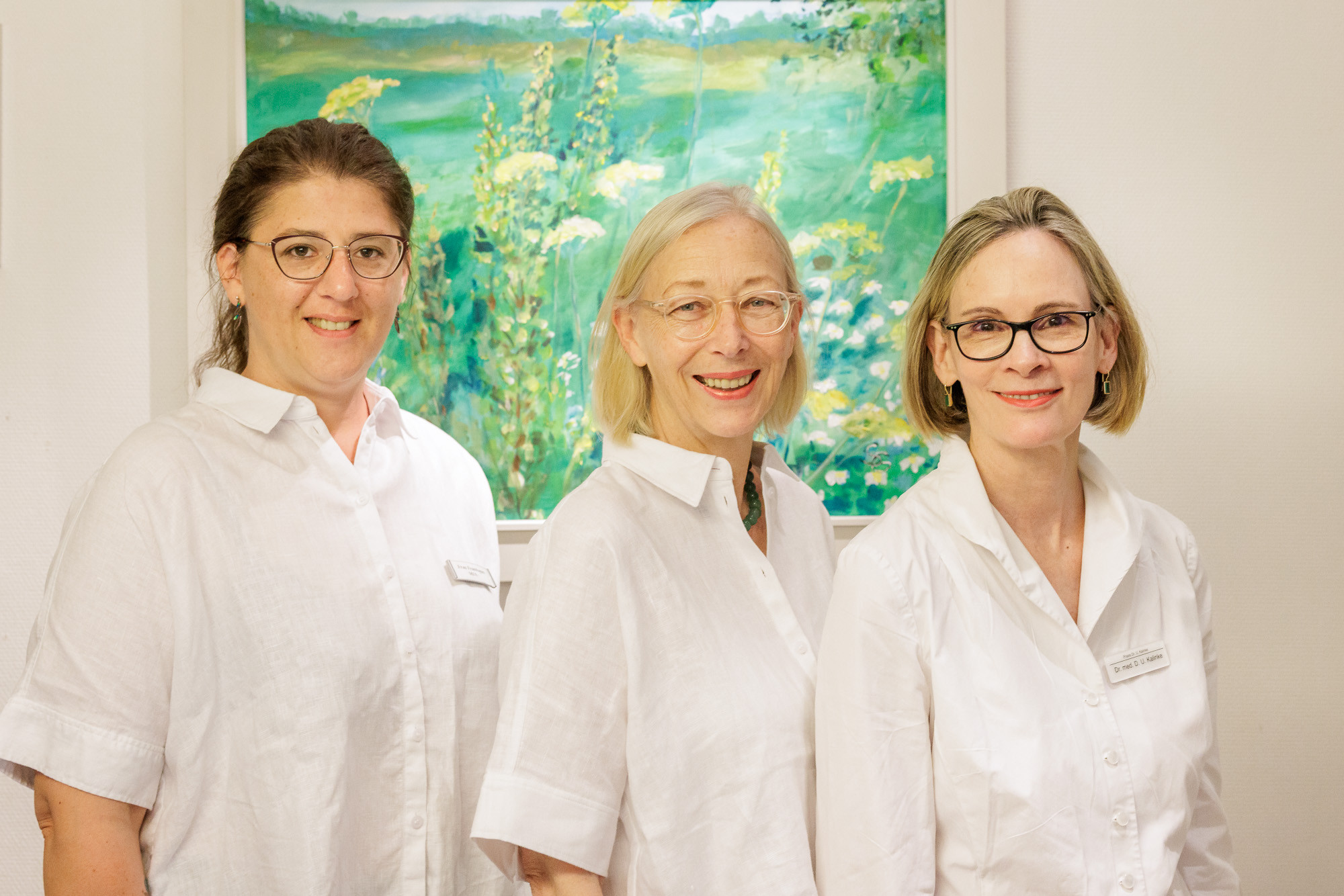 Teamfoto aus einer Arztpraxis mit drei Mitarbeiterinnen in weißer Kleidung vor einem grünen Gemälde