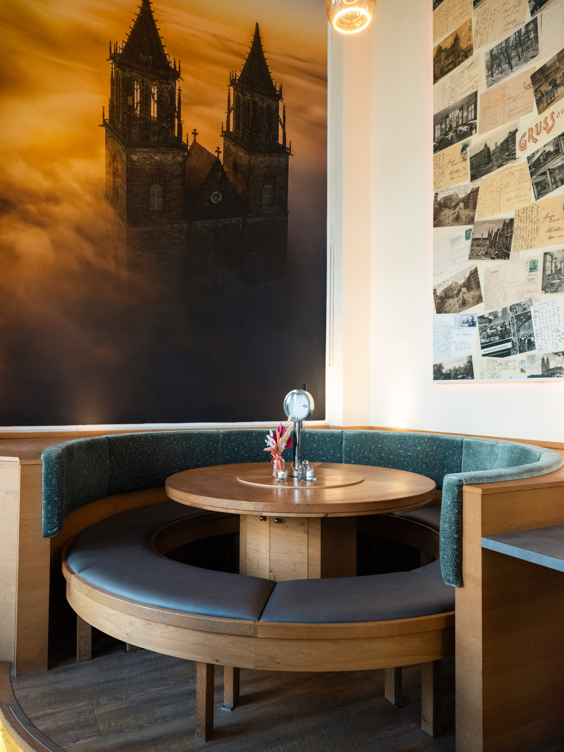 Runder Sitzbereich in einem Café mit Holztisch, Wandbild einer Kirche im Nebel und alten Postkarten an der Wand