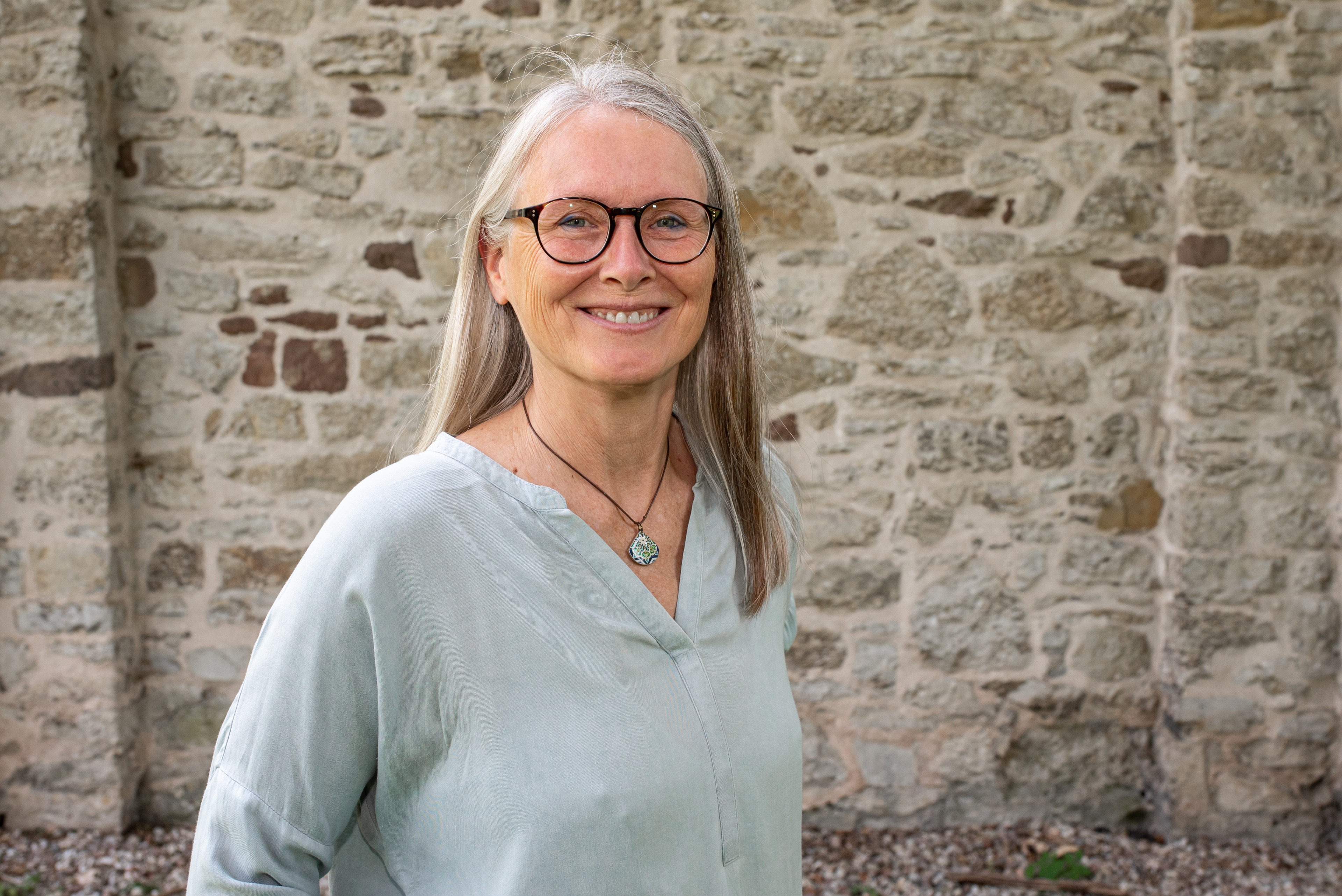 Verena vor einer Steinmauer mit hellblauer Bluse und Kette