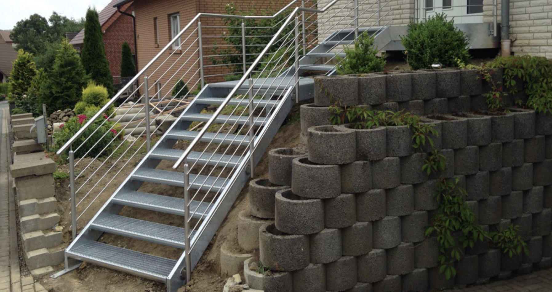 Außentreppe aus Metall mit Edelstahlgeländer an einer Hausfassade, neben einer Mauer aus Betonsteinen