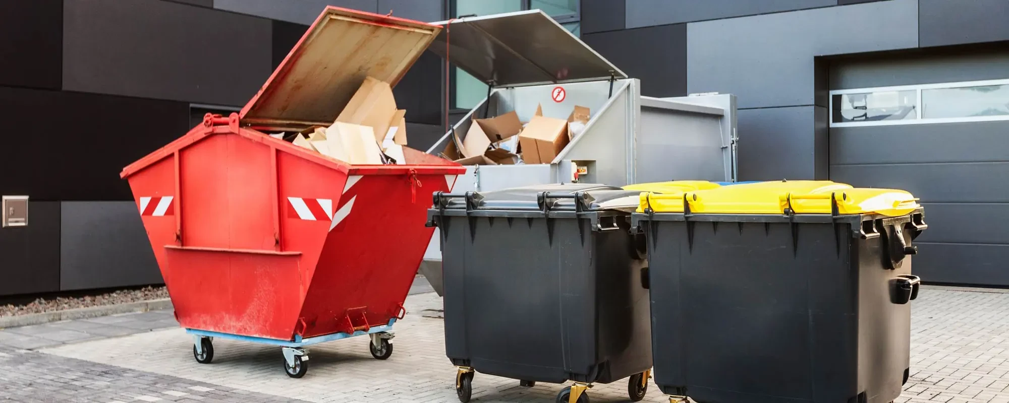 Verschiedene Müllcontainer und Abfallbehälter vor einem Gebäude zur Entsorgung von Karton und Müll
