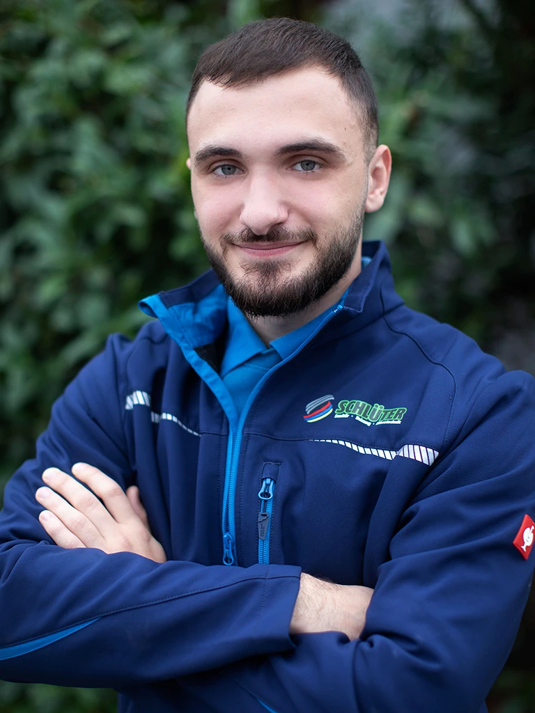 Enis Marisenovic in Schlüter Arbeitskleidung mit verschränkten Armen vor grüner Natur