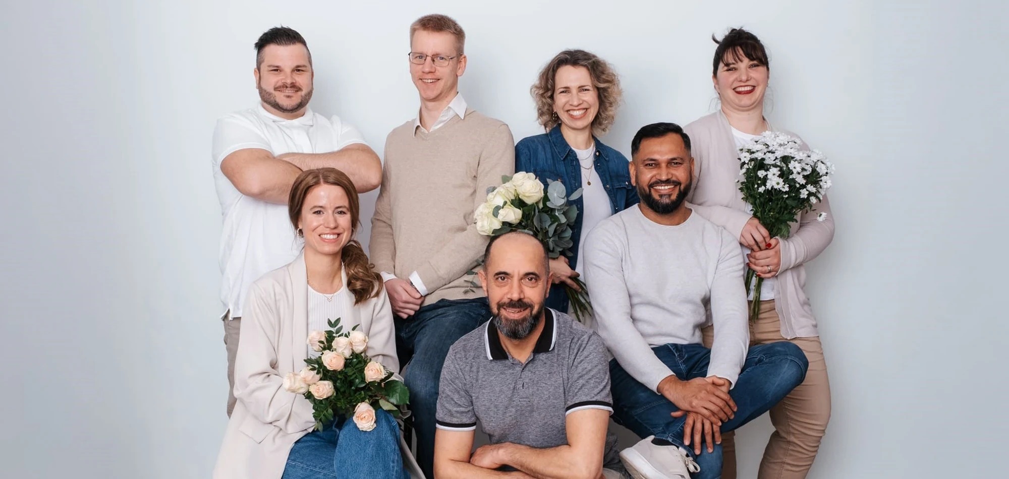Neues Teamfoto einer Gruppe von Mitarbeitern mit Blumen, sitzend und stehend vor hellem Hintergrund