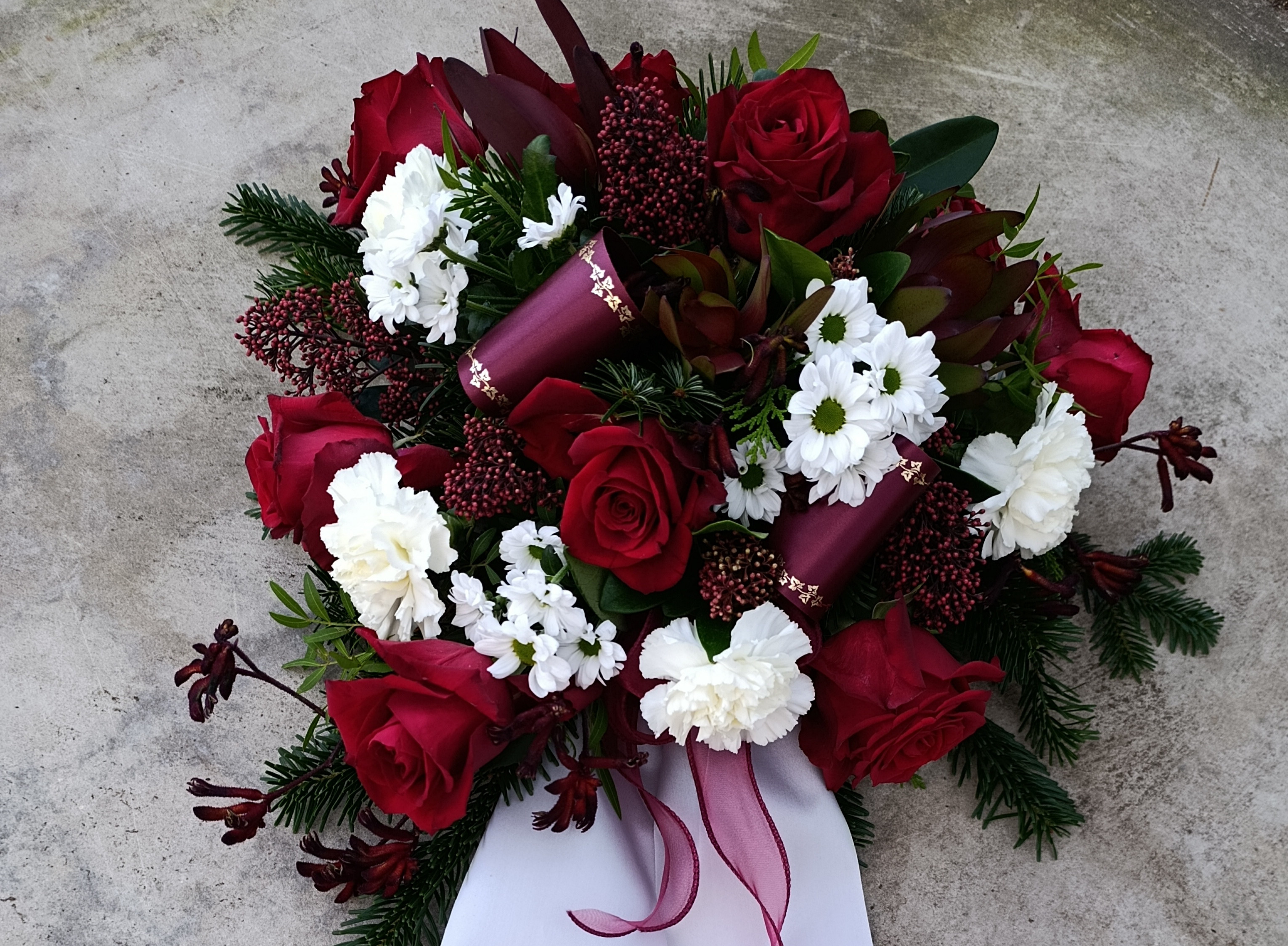 Trauergesteck mit roten Rosen und weißen Nelken auf Steinboden, dekoriert mit Schleife