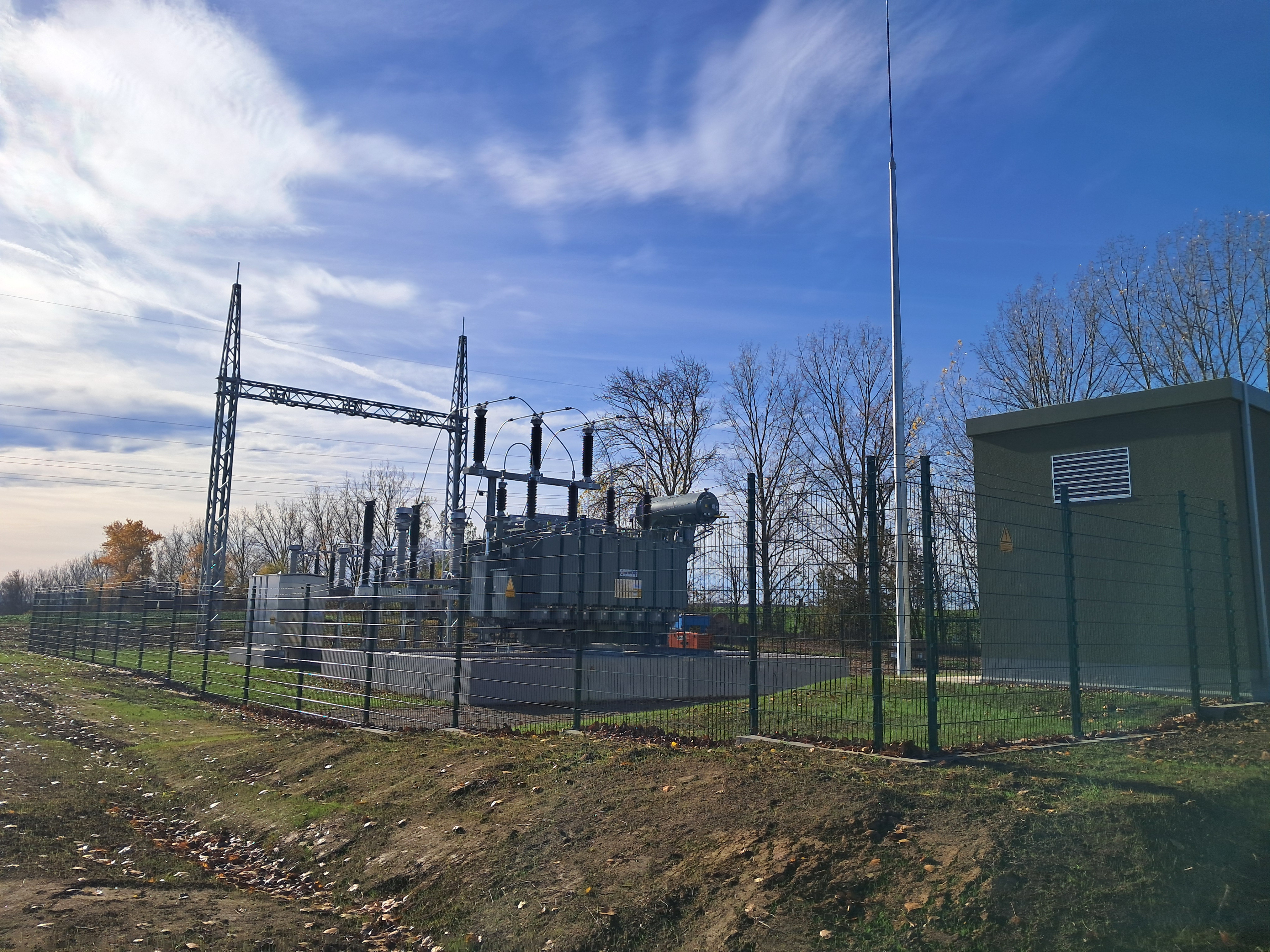 Umspannwerk mit Hochspannungsanlage und Transformator im Freien, umgeben von Zaun und Bäumen