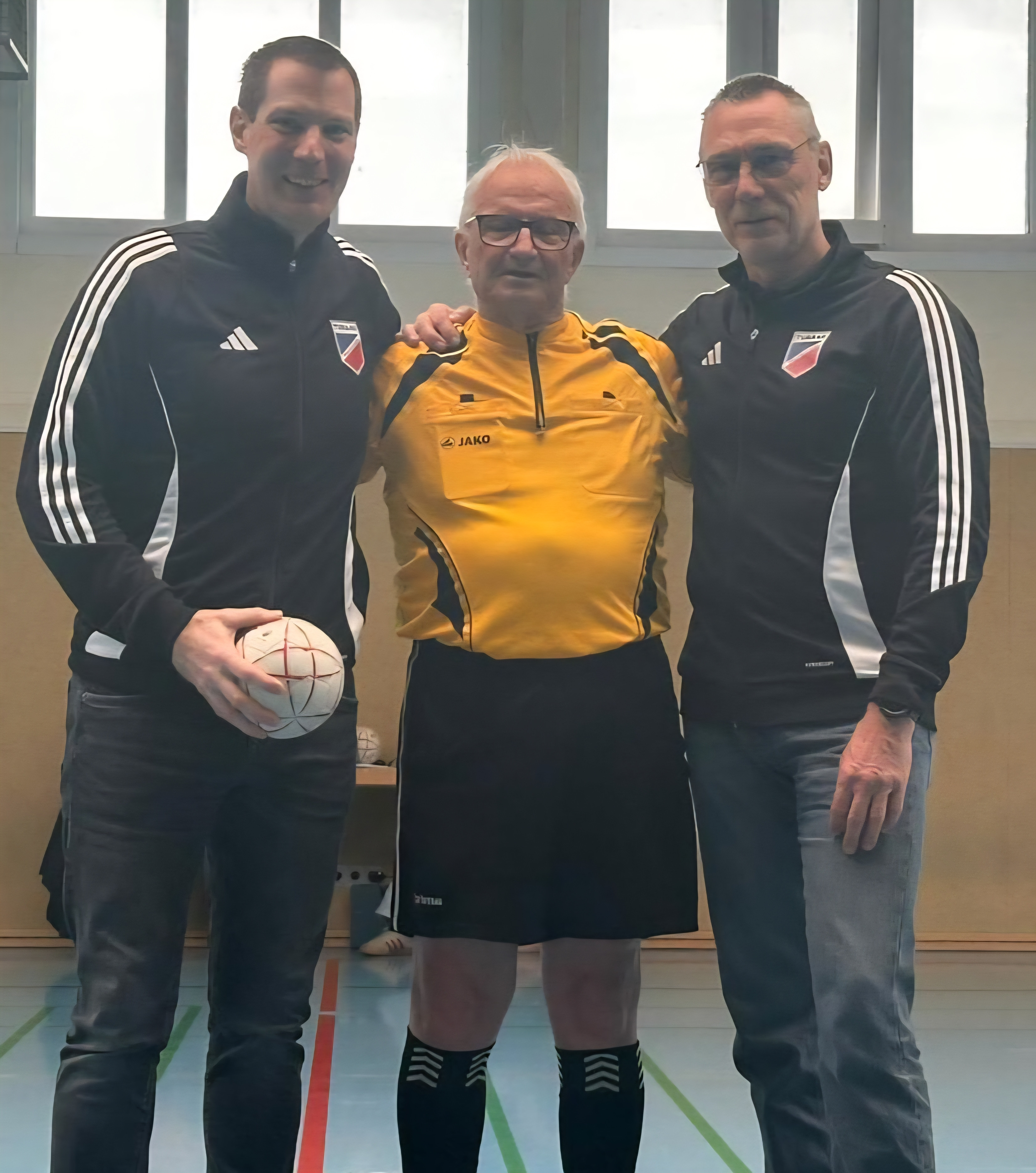 Drei Männer in Sportkleidung in einer Sporthalle, zwei mit Vereinsjacken und einer als Schiedsrichter, Hallensportball in der Hand