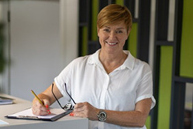Petra Dammann 2025 – Porträtaufnahme mit Klemmbrett und Brille Petra Dammann im Jahr 2025 mit weißem Hemd und Brille, schreibt auf einem Klemmbrett