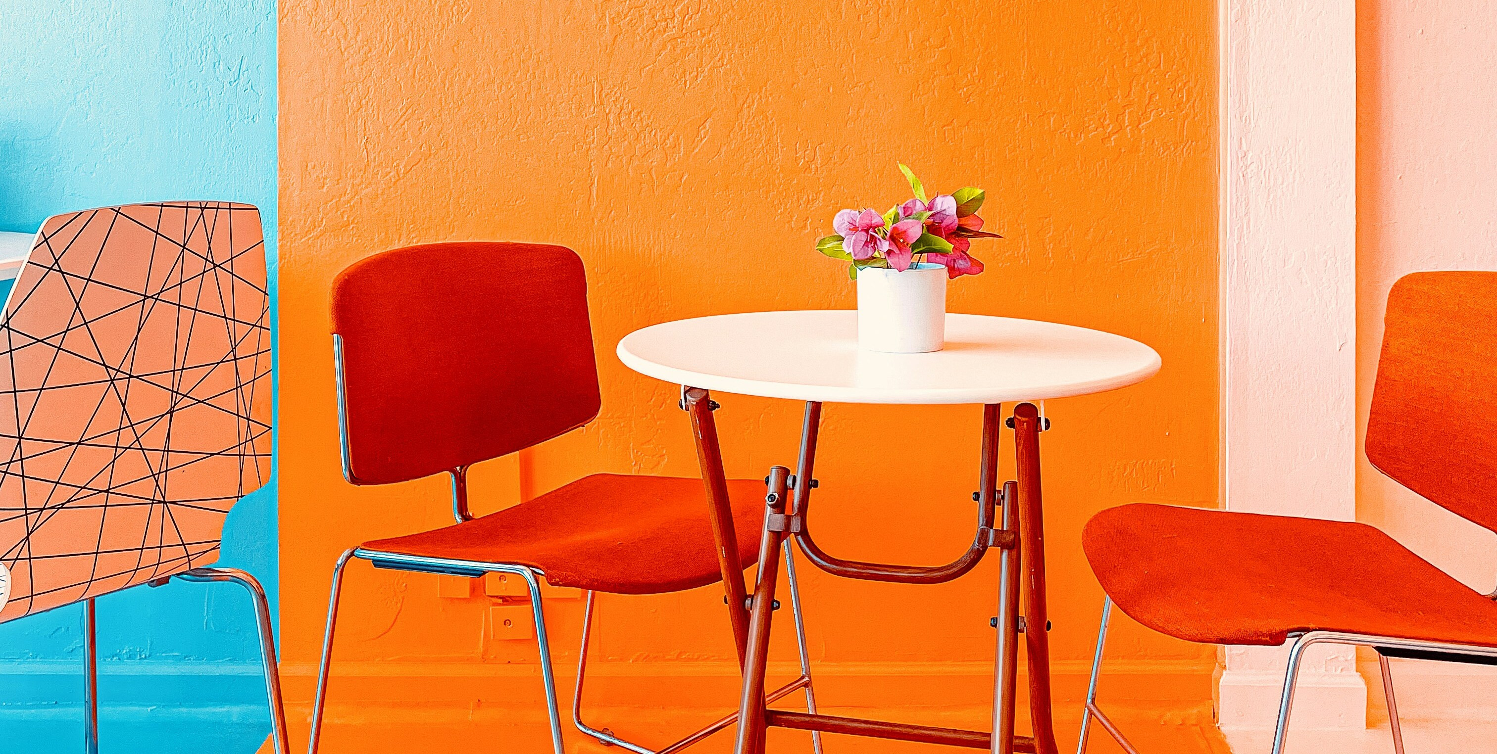 Knalliges Farbkonzept mit orangefarbener Wand, roten Stühlen und weißem Tisch mit Blumen
