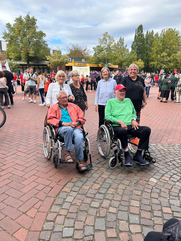Menschen in Rollstühlen und Begleiter bei einer öffentlichen Veranstaltung auf einem Platz Gruppe von Menschen, darunter zwei Personen im Rollstuhl, bei einer Veranstaltung im Freien auf einem Platz mit vielen weiteren Teilnehmern im Hintergrund.