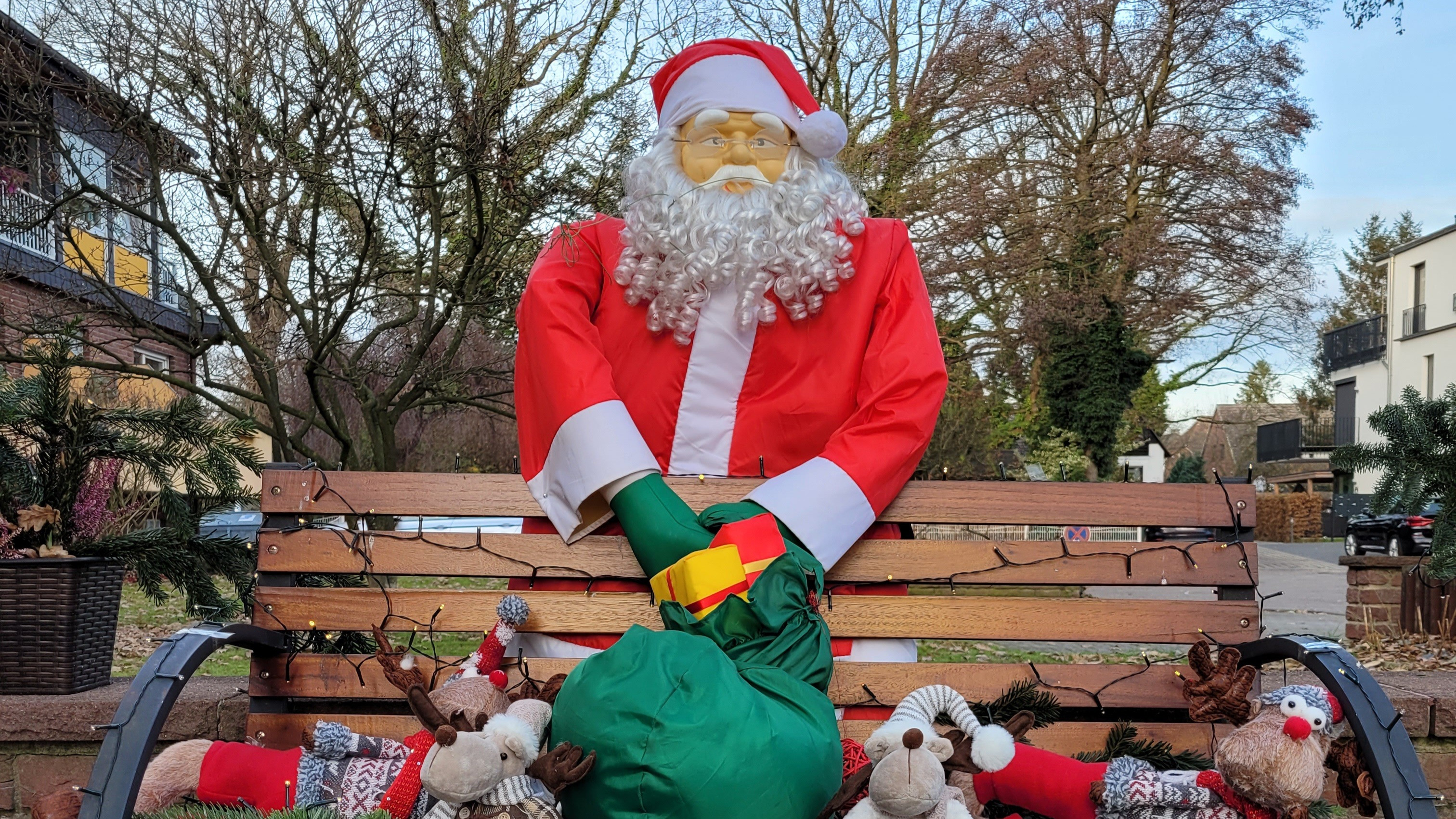 Weihnachtsmann in rotem Kostüm mit Geschenkesack und Rentier-Plüschtieren auf einer Bank im Freien
