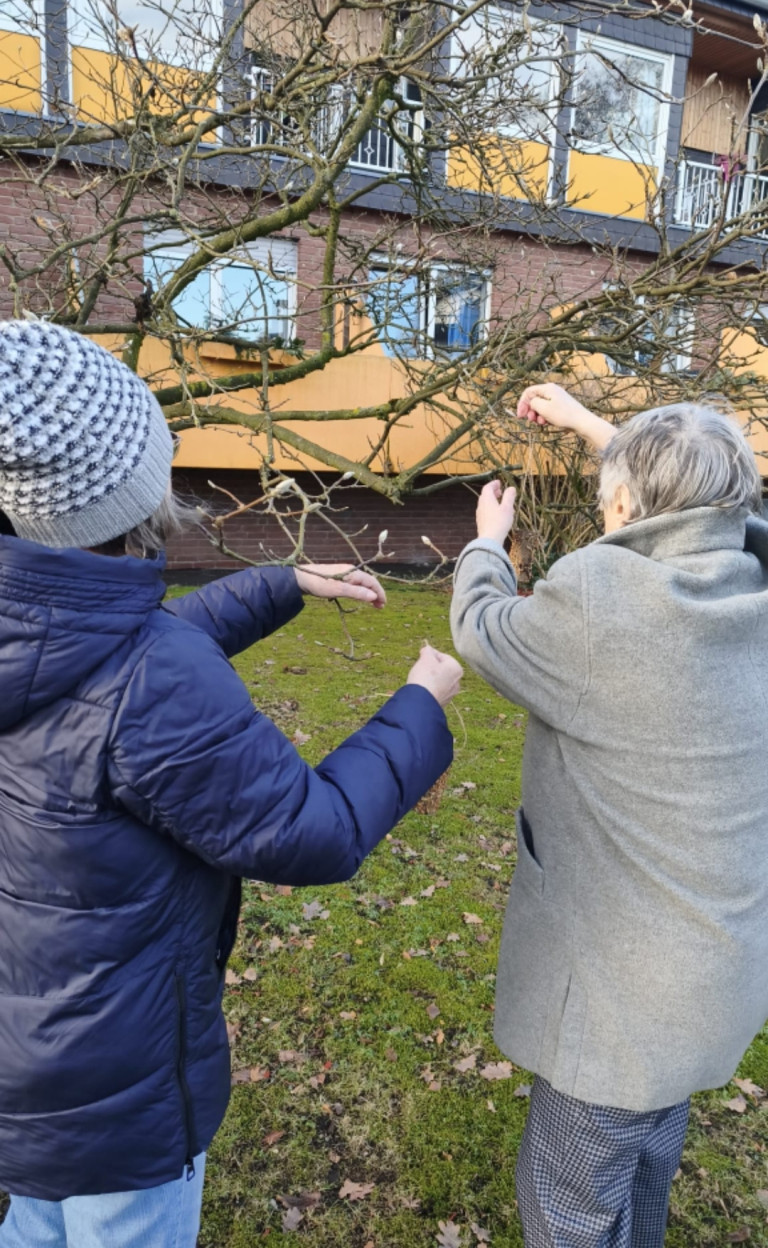 Zwei ältere Frauen hängen gemeinsam Dekoration an einen Baum im Garten vor einem Haus
