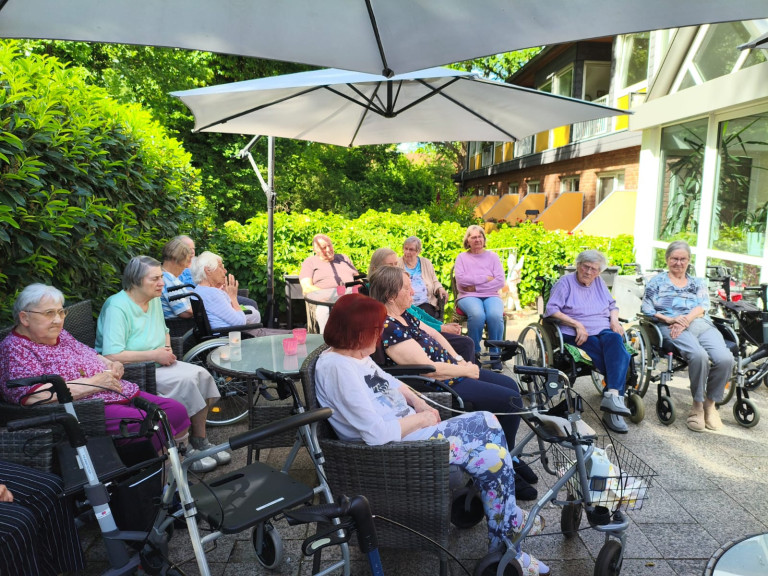 Senioren sitzen gemeinsam im Garten eines Pflegeheims unter einem Sonnenschirm und genießen das schöne Wetter.