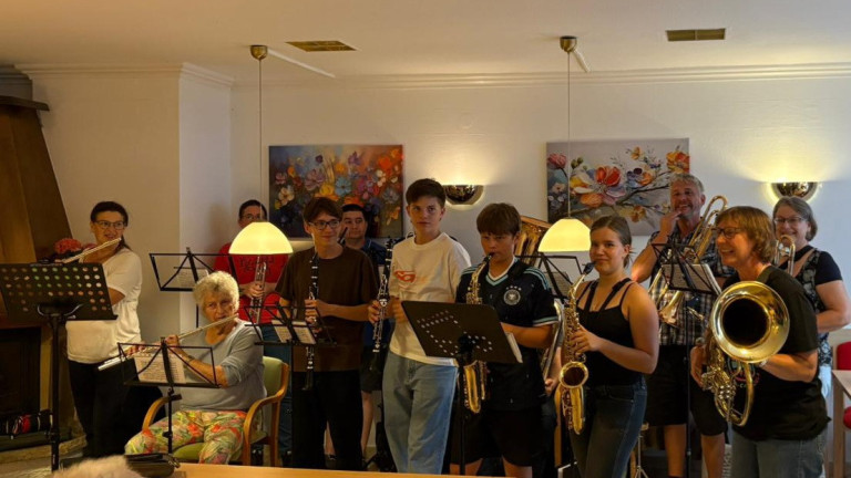 Gemeinsames Musizieren von Jugendlichen und Senioren mit Blasinstrumenten am 28.08.2026 Jugendliche und Senioren musizieren gemeinsam mit Blasinstrumenten in einem Wohnzimmer, Gruppenfoto mit Notenständern und Gemälden im Hintergrund.