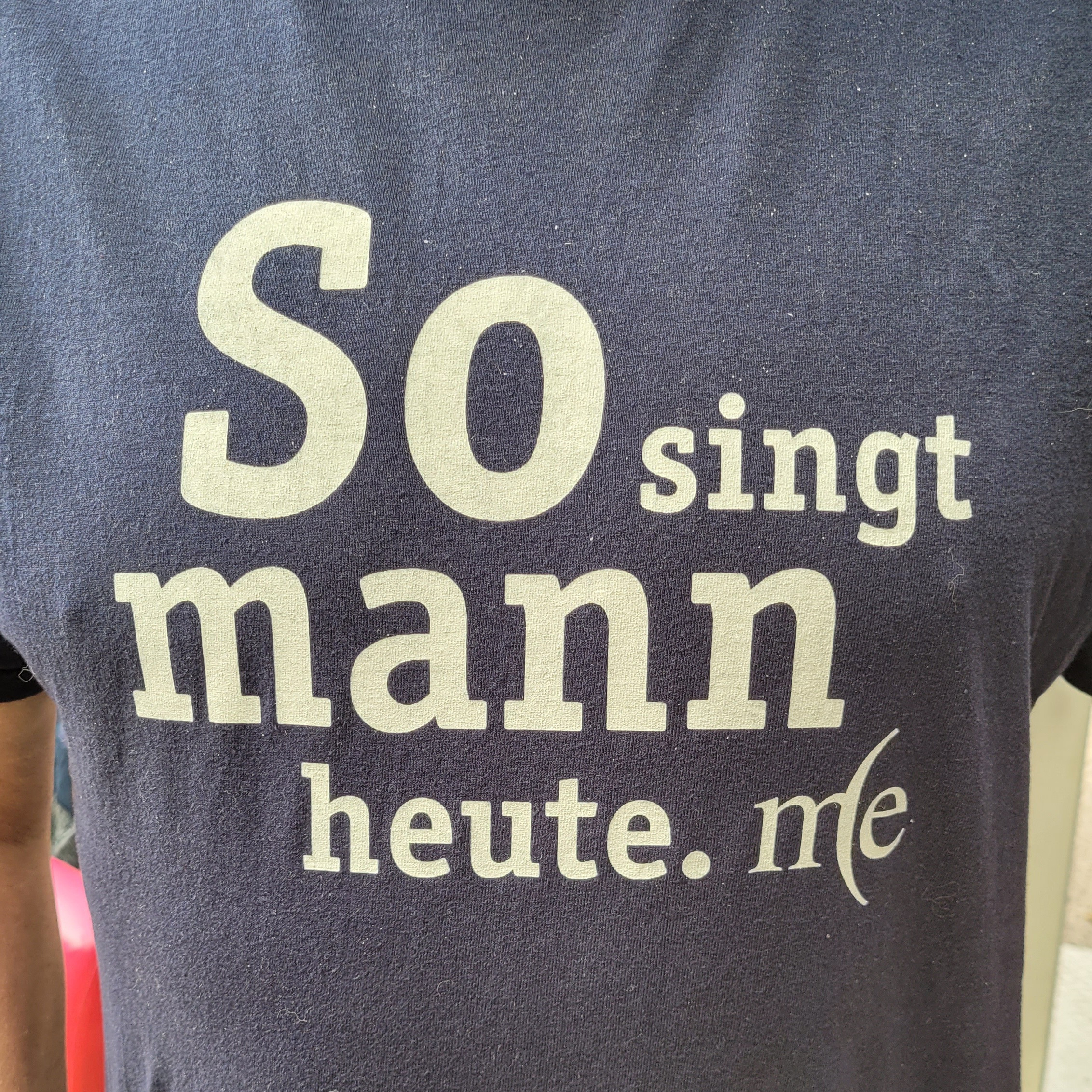 T-Shirt mit Aufdruck 'So singt mann heute. me' in weißer Schrift auf dunklem Stoff