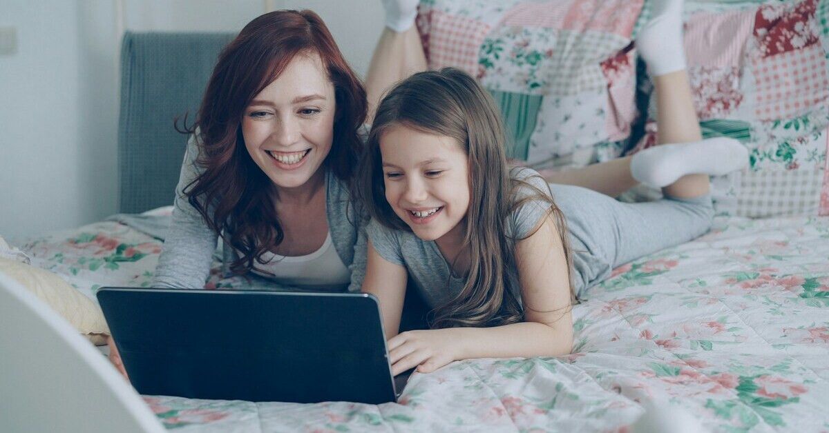 Mutter und Kind lachen zusammen auf einem Bett und schauen auf ein Tablet.