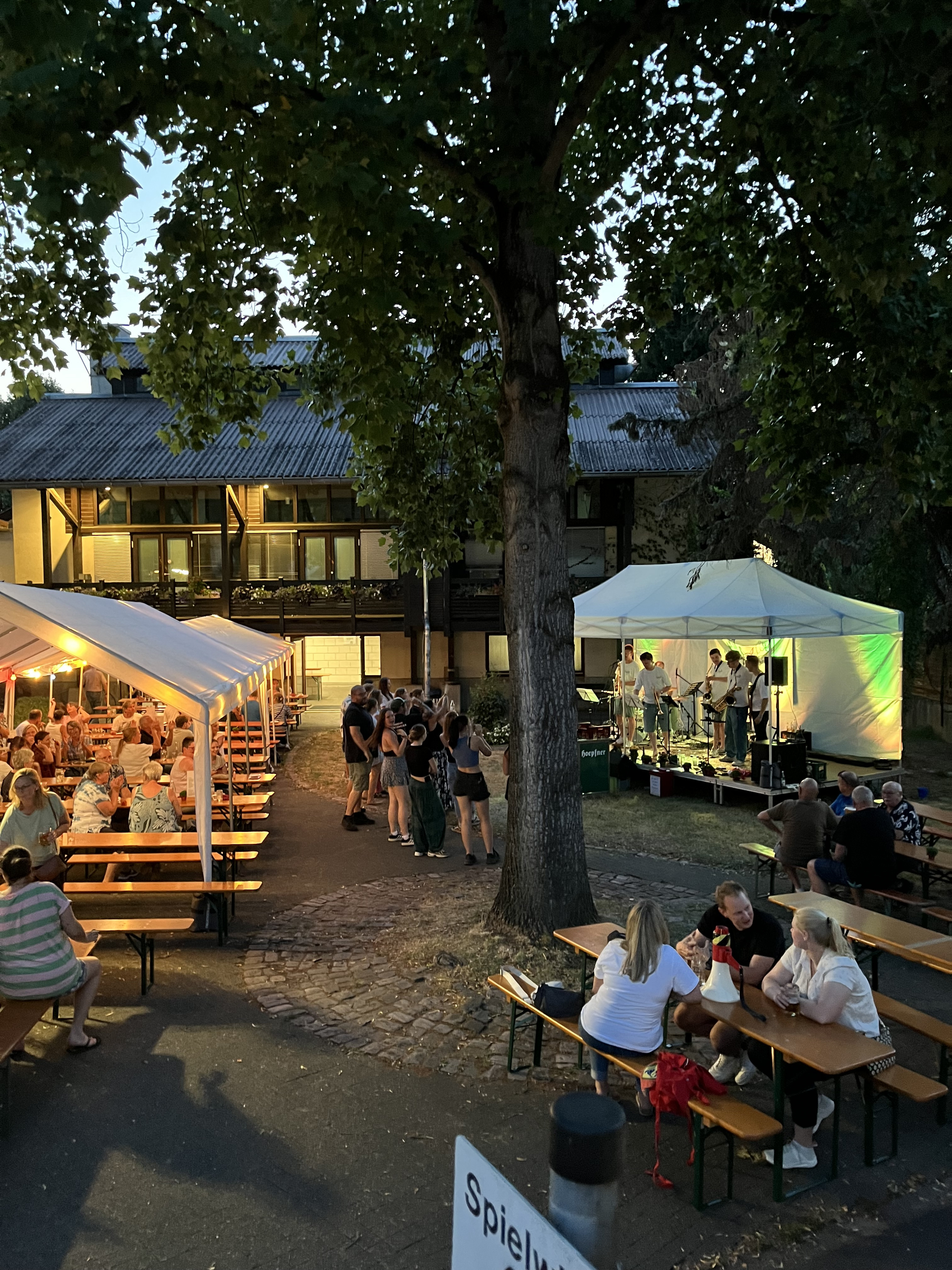 Abendliches Sommerfest mit Live-Band, Menschen an Bierzeltgarnituren und Pavillon im Freien