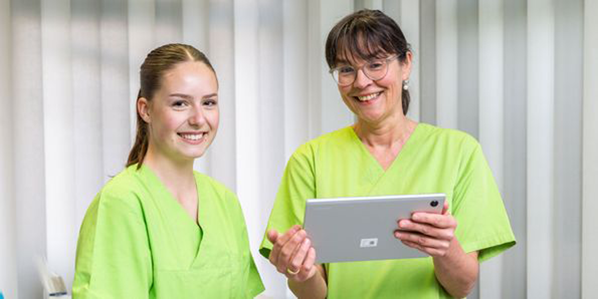 Zwei Pflegekräfte in grüner Arbeitskleidung betrachten ein Tablet im medizinischen Umfeld