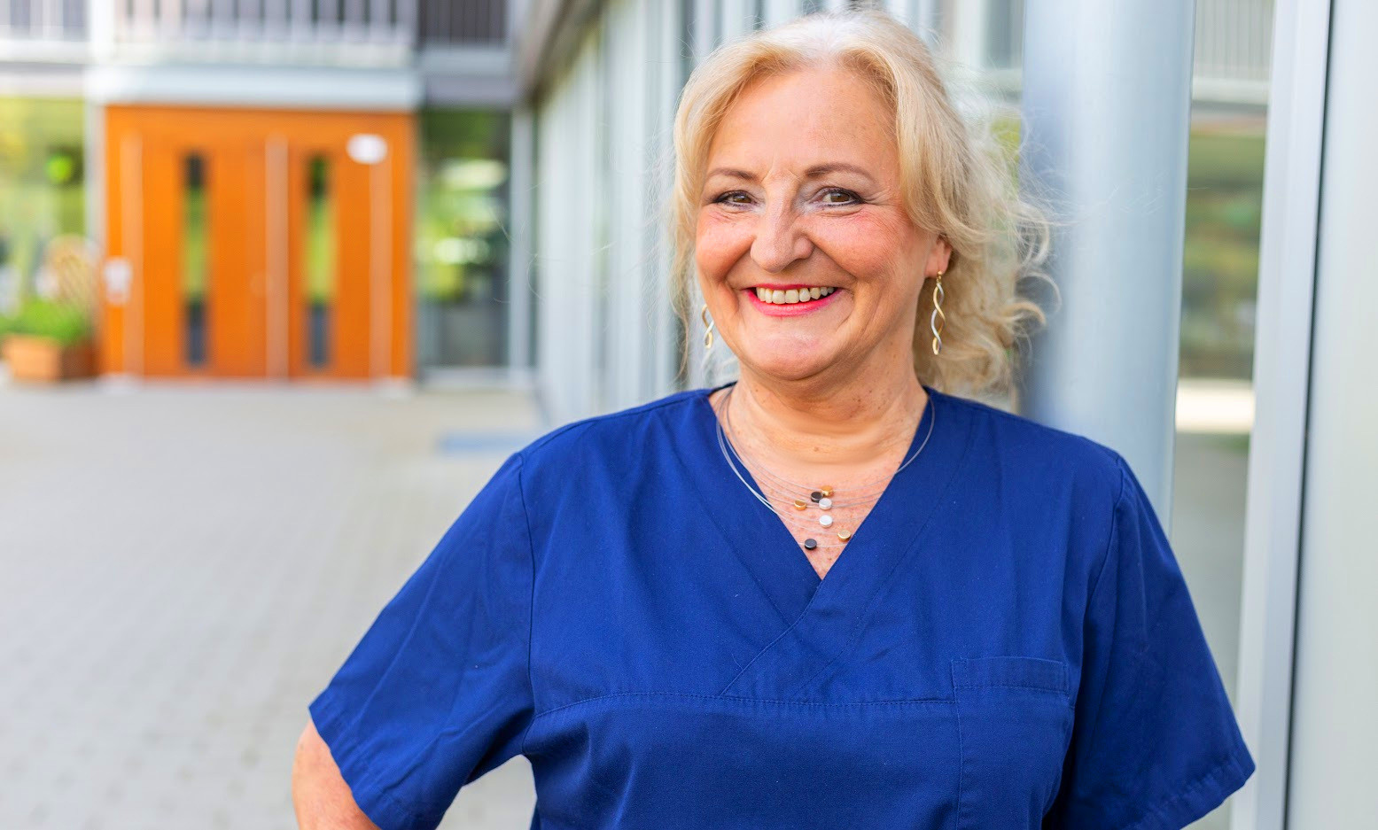 Ute in blauer Pflegekleidung vor einem modernen Gebäude