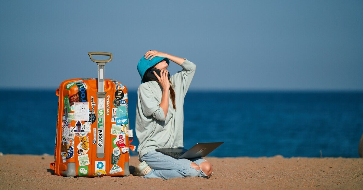 Frau mit Laptop und Handy am Strand, gestresst im Urlaub neben Koffer