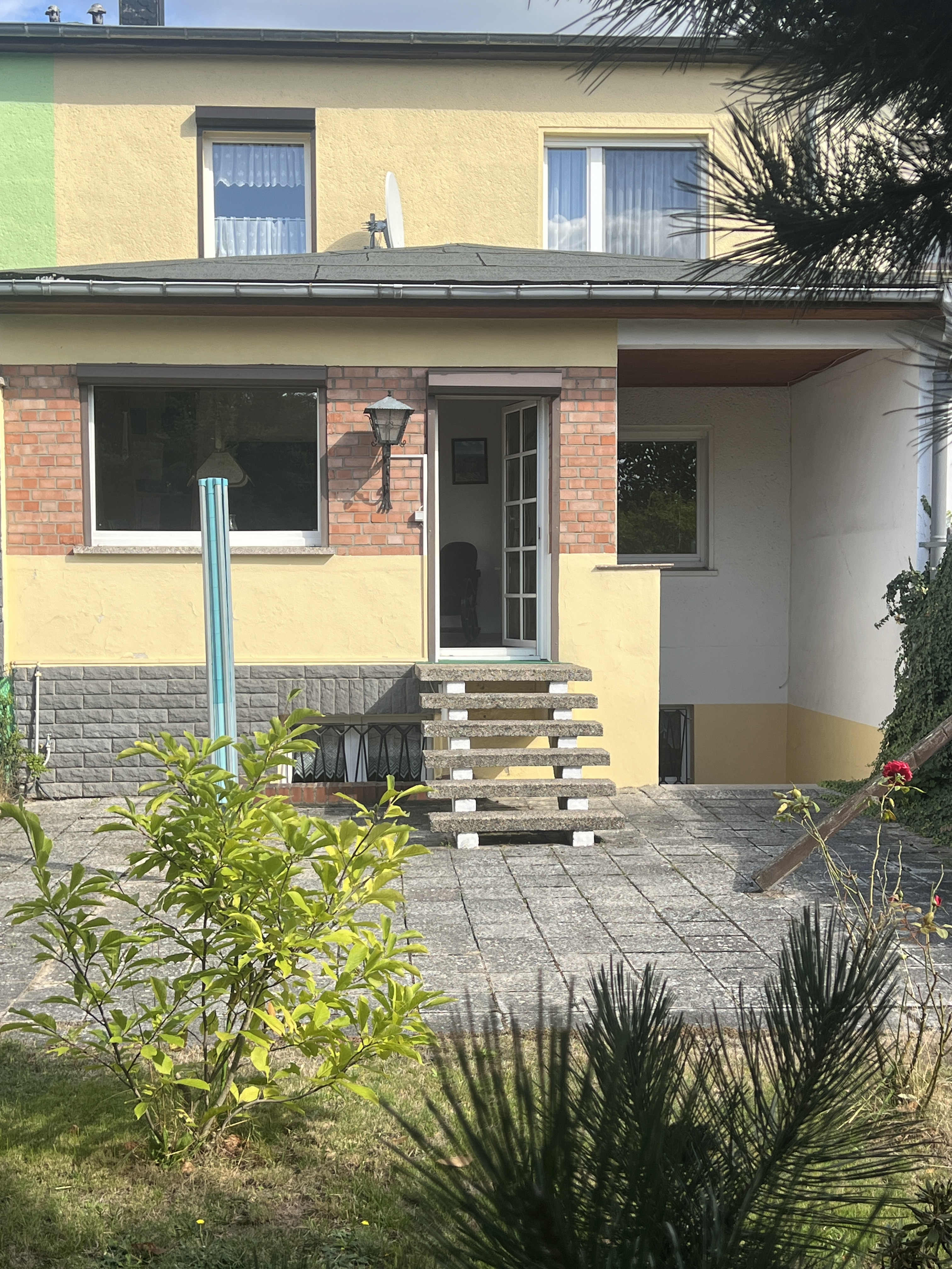 Hauseingang mit Treppe, Terrasse und Gartenpflanzen an einem sonnigen Tag