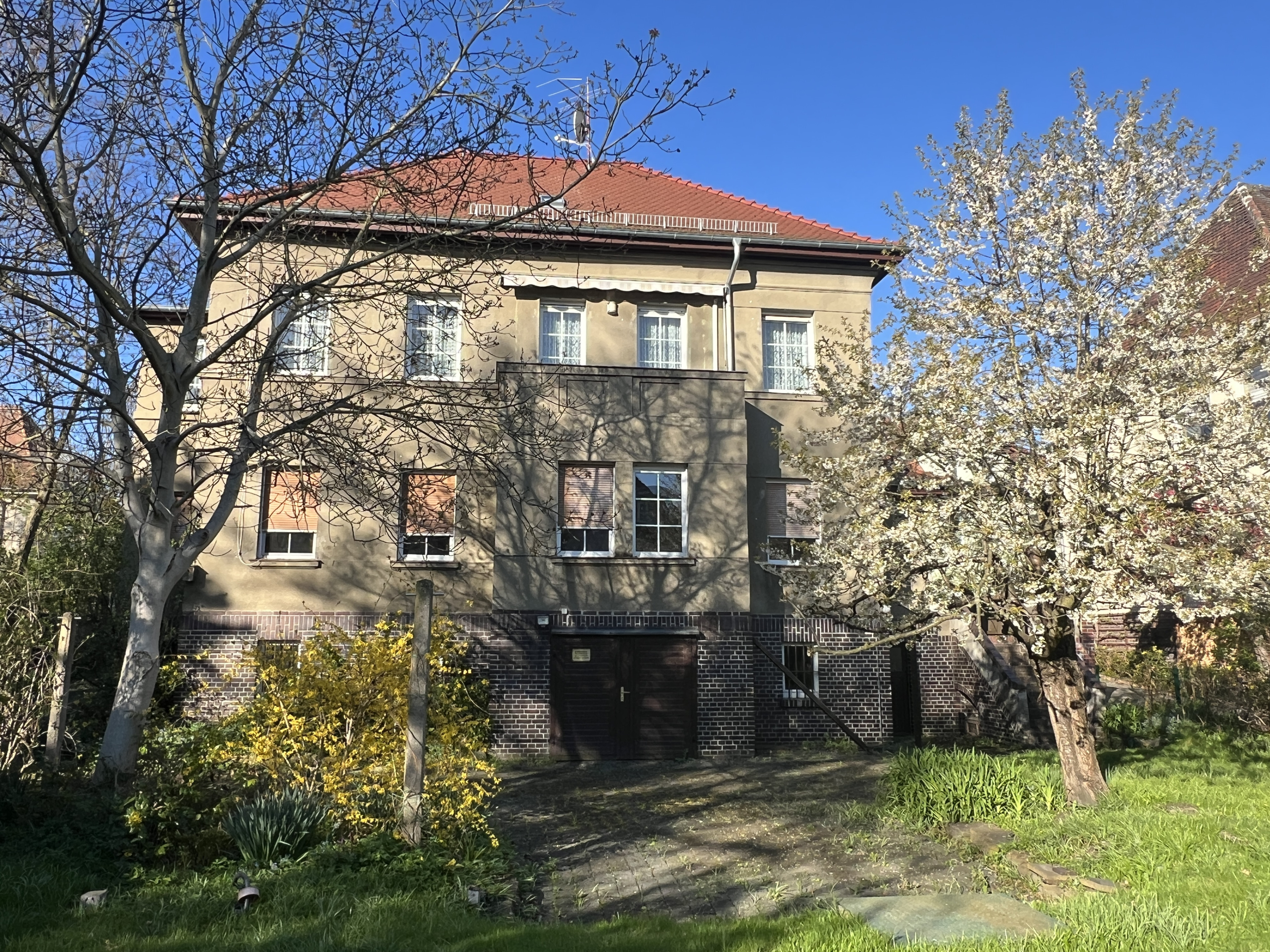 Mehrfamilienhaus mit rotem Dach und blühenden Bäumen im Frühling vor blauem Himmel