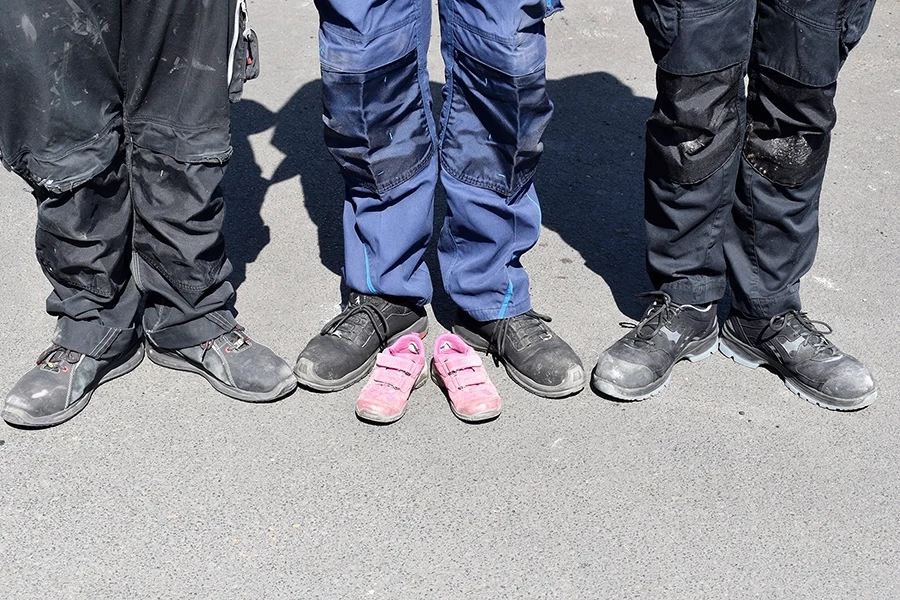 Beine von Arbeitern in Sicherheitsschuhen mit kleinen pinken Schuhen dazwischen