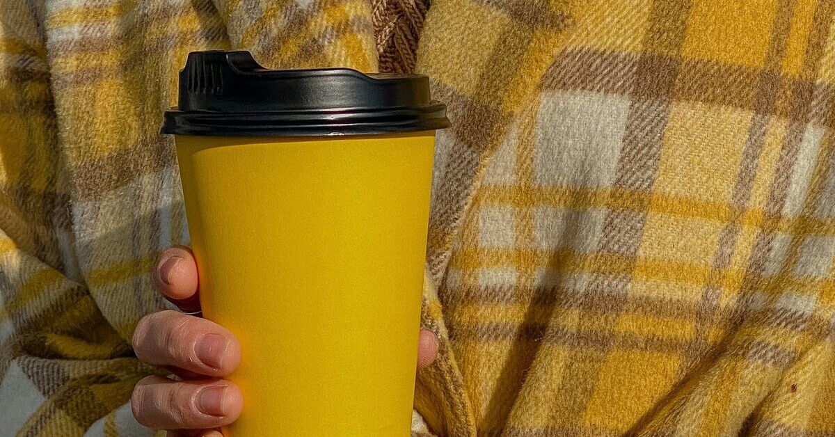Coffee to go Becher in gelb wird in der Hand gehalten, im Hintergrund eine karierte Jacke
