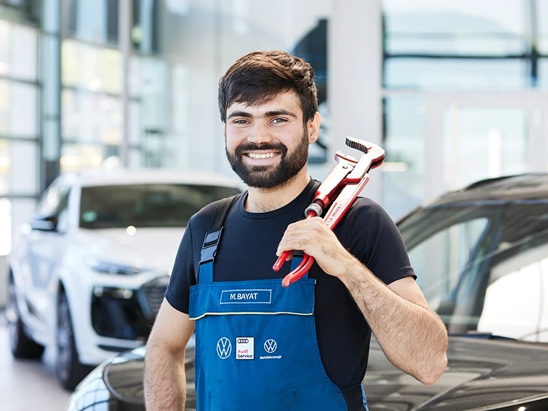 Mohammad Bayat als Kfz-Mechaniker im Autohaus mit Werkzeug vor einem VW und Audi Fahrzeug