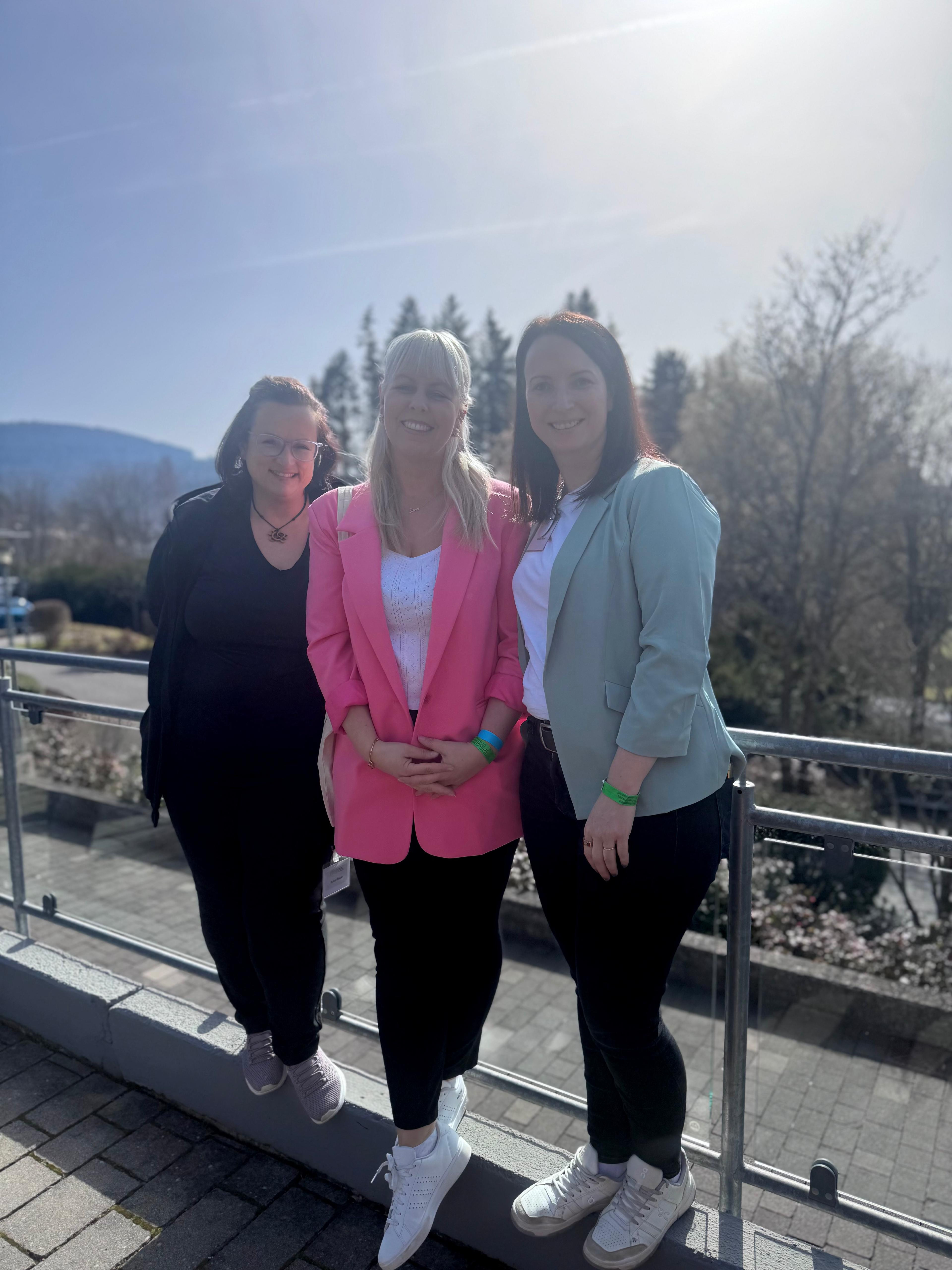 Drei Frauen im Business-Outfit stehen draußen auf einer Terrasse mit Bäumen und Bergen im Hintergrund