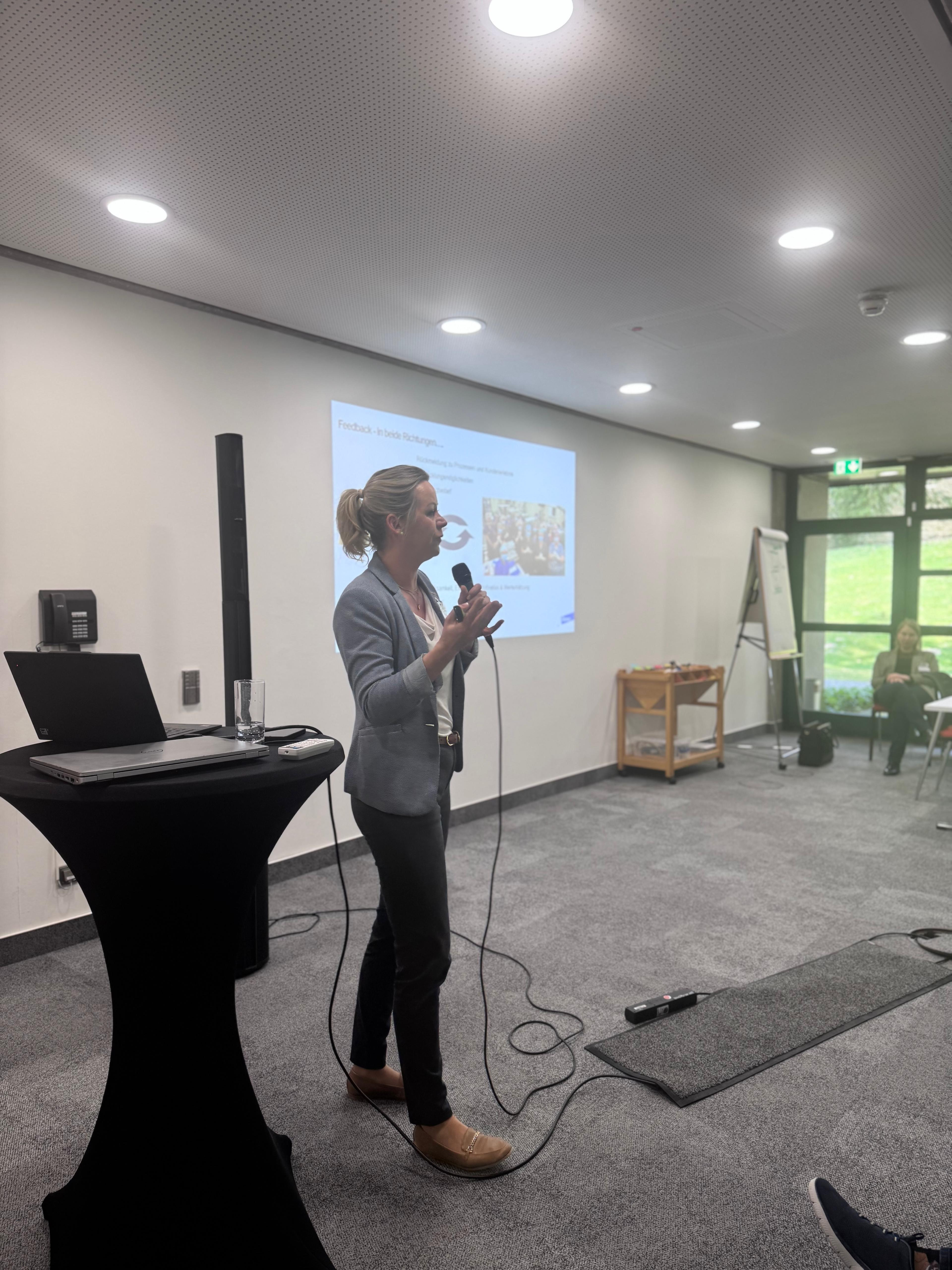 Frau hält Präsentation mit Mikrofon in modernem Seminarraum vor Projektionsfläche
