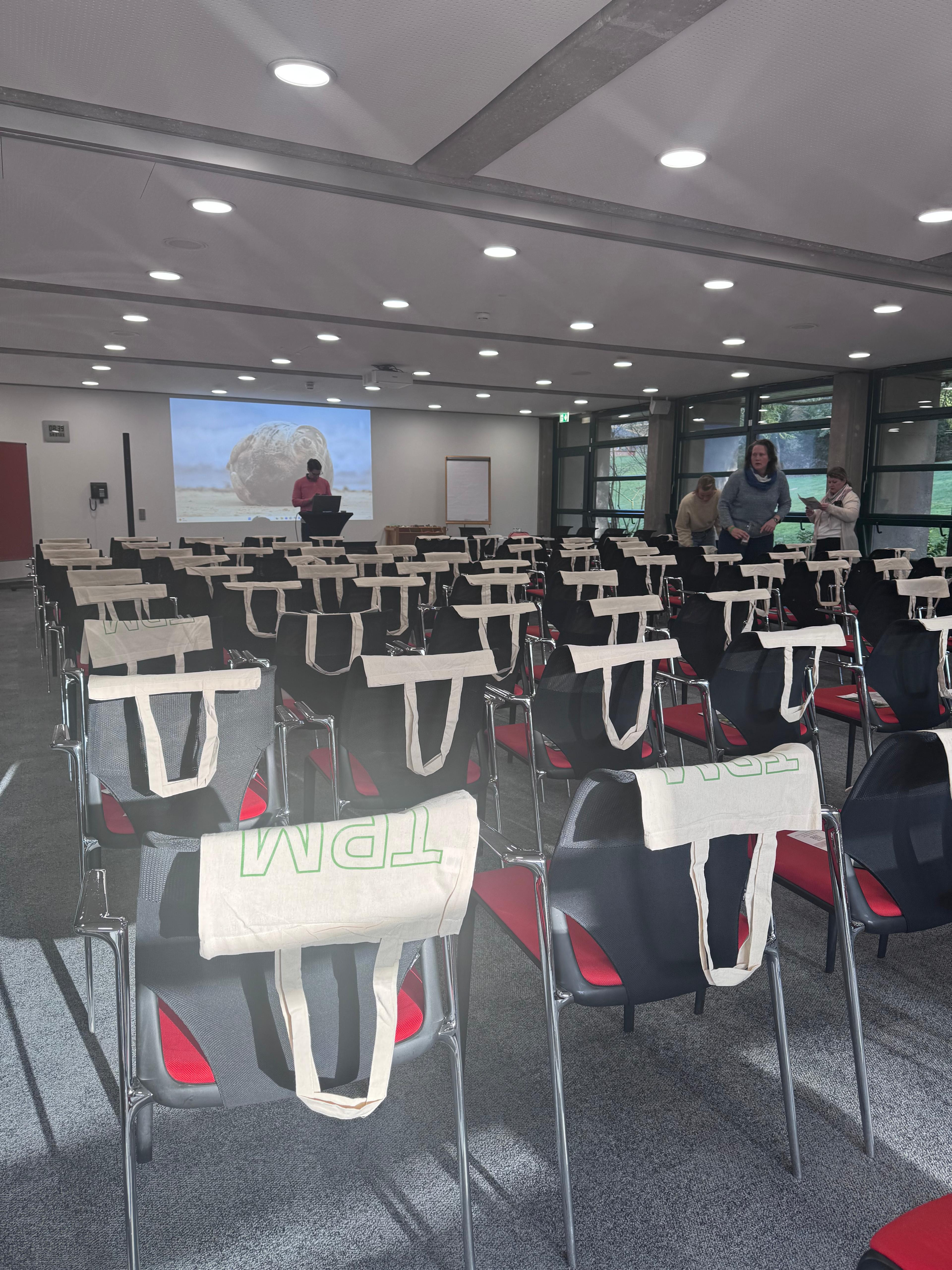 Konferenzraum mit vorbereiteten Stühlen und Stofftaschen, Präsentation und Teilnehmer im Hintergrund