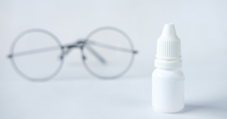 Weiße Flasche mit Augentropfen vor unscharfer Brille auf weißem Hintergrund