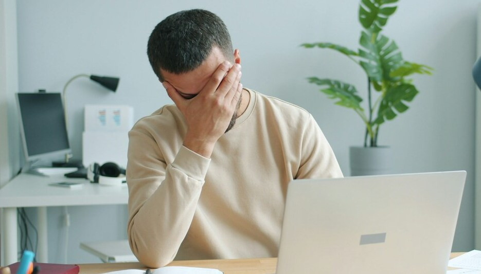 Angestrengt und belastet: Gestresster Mann am Arbeitsplatz mit Laptop Angestrengt und belastet wirkender Mann am Schreibtisch mit Laptop, der sich gestresst das Gesicht h