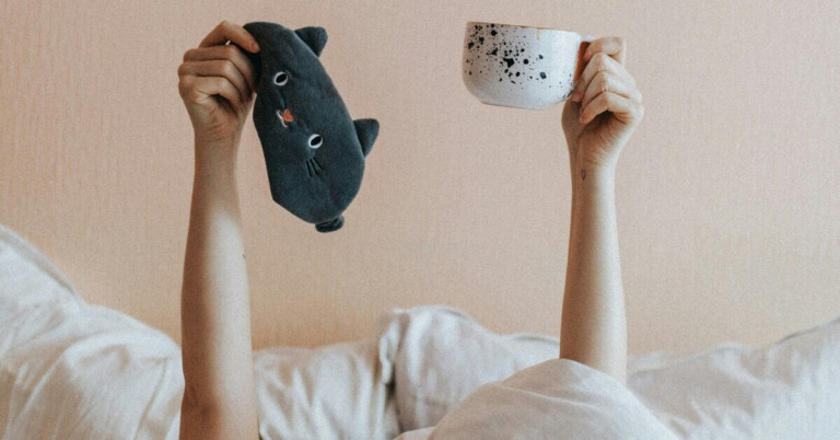 Kaffee vorm Schlafen – Person hält eine Tasse Kaffee und eine Schlafmaske im Bett