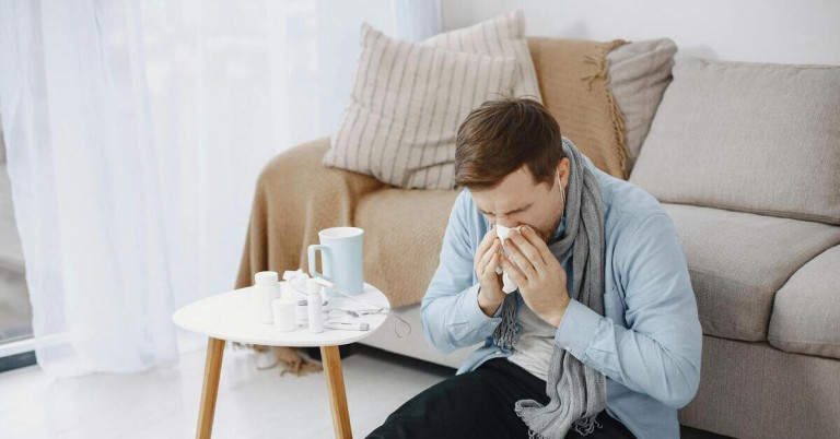 Mann mit Erkältung sitzt auf dem Sofa und benutzt ein Taschentuch, Medikamente und Tee auf dem Tisch