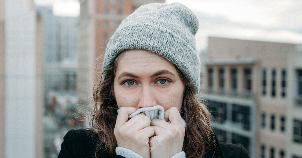 Frau mit Mütze hält sich frierend die Hände vor das Gesicht, Symbolbild für Erkältung