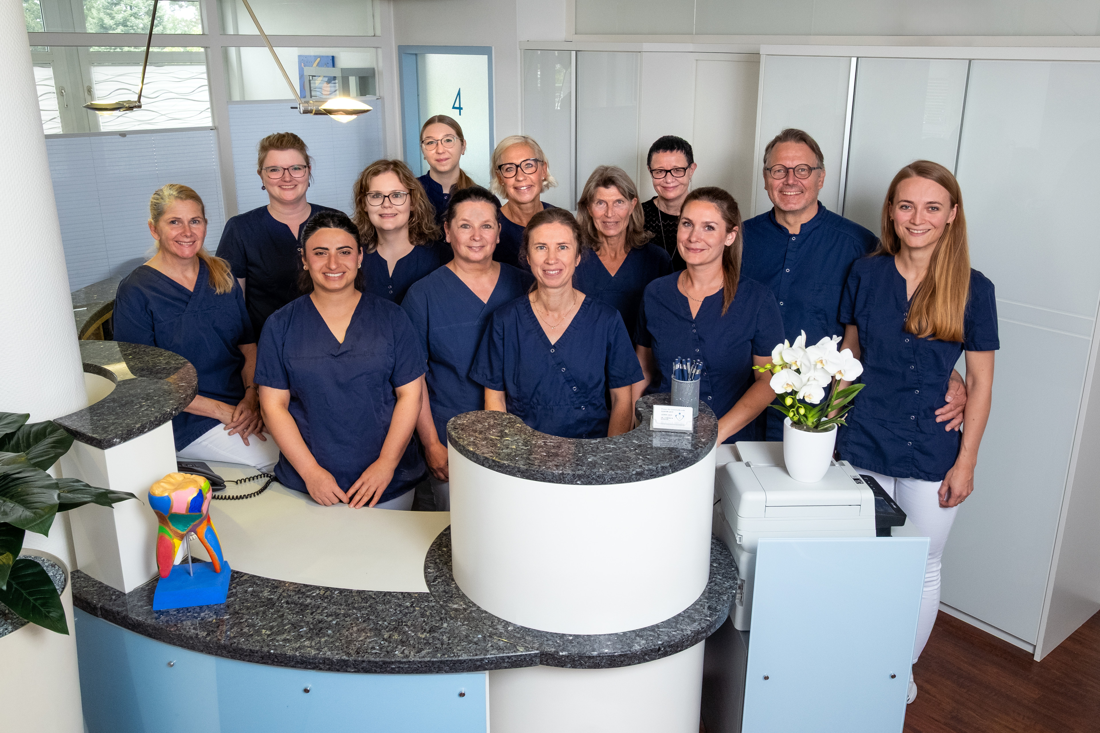 Gruppenfoto des Praxisteams am Empfangstresen in blauer Arbeitskleidung Gruppenfoto eines Praxisteams in blauer Arbeitskleidung an einem Empfangstresen