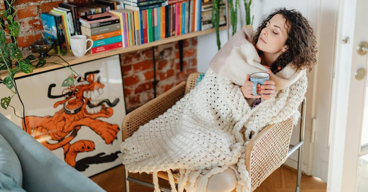 Frau friert auf Sofa, eingekuschelt in Decke und hält eine Tasse Tee im Wohnzimmer