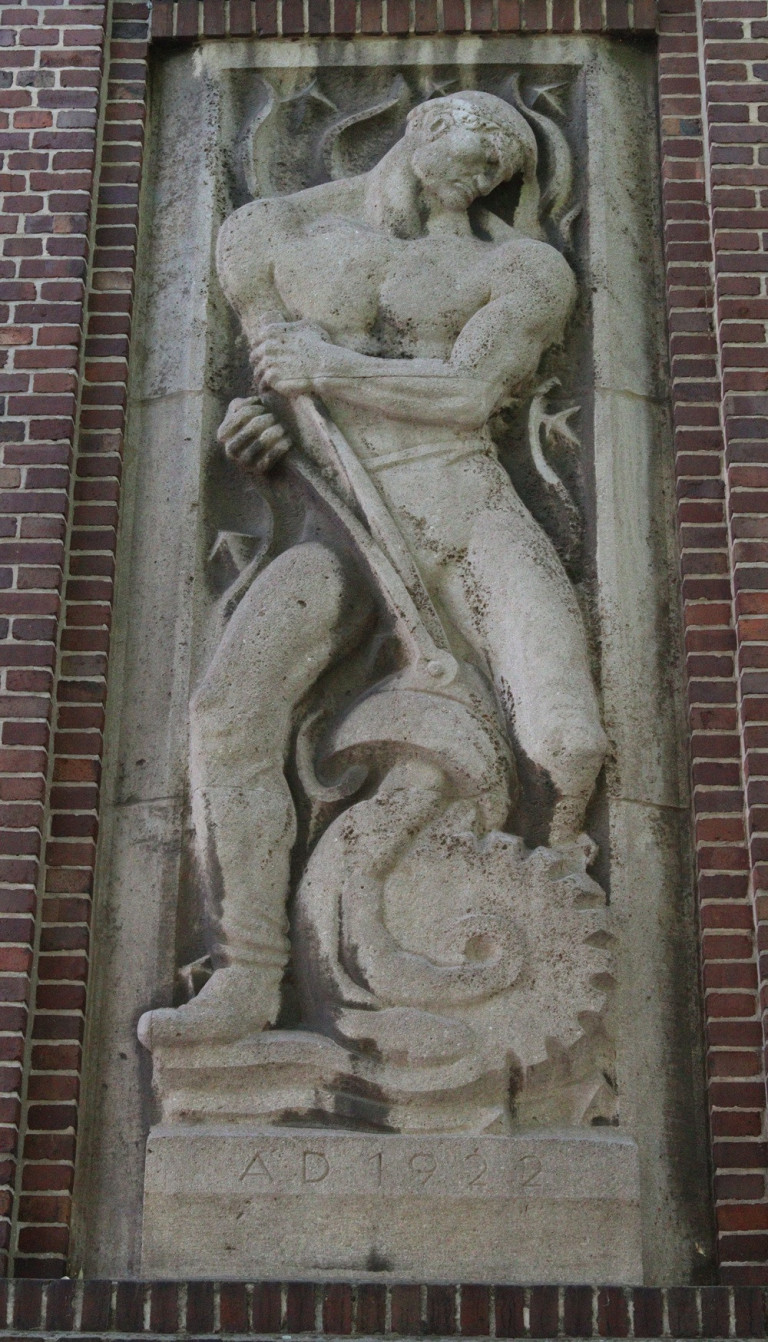 Ausschnitt eines Reliefs mit einer muskulösen Figur, die ein Werkzeug hält, vor einer Backsteinwand