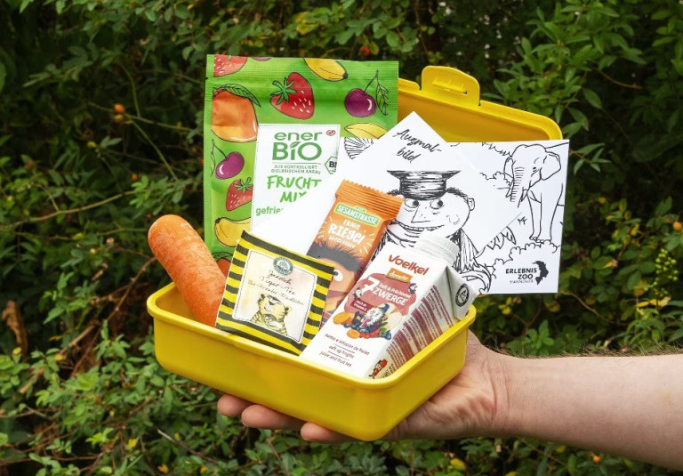 Biobrotbox 2025 mit gesunden Snacks, Karotte, Bio-Fruchtsnack, Müsliriegel, Getränk und Ausmalbild in gelber Brotdose