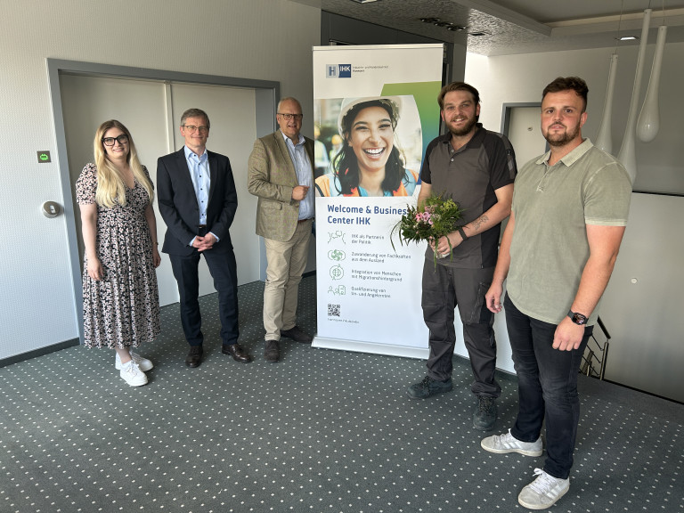 Anerkennungspartnerschaft im Welcome & Business Center IHK – Gruppenfoto bei offizieller Veranstaltung Gruppenfoto bei einer Veranstaltung zur Anerkennungspartnerschaft im Welcome & Business Center IHK, mit Personen und Blumenstrauß vor einem IHK-Banner.