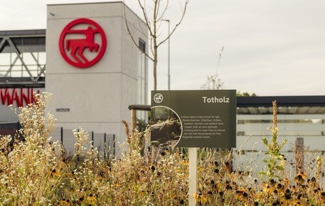 Totholz-Infotafel vor moderner Firmenzentrale mit rotem Tier-Logo, naturnahe Bepflanzung im Vordergrund