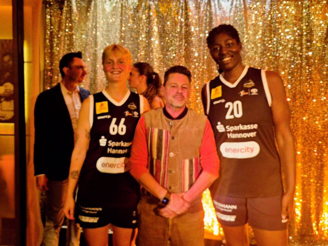 Zwei Basketballspielerinnen vom Team Sparkasse Hannover mit Trikots und einer weiteren Person vor goldenem Hintergrund