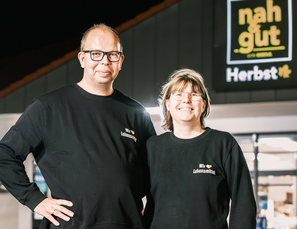 Zwei Personen in schwarzer Kleidung mit Aufdruck 'Wir lieben Lebensmittel' vor einem Supermarkt, Thema Tüte und Tonne